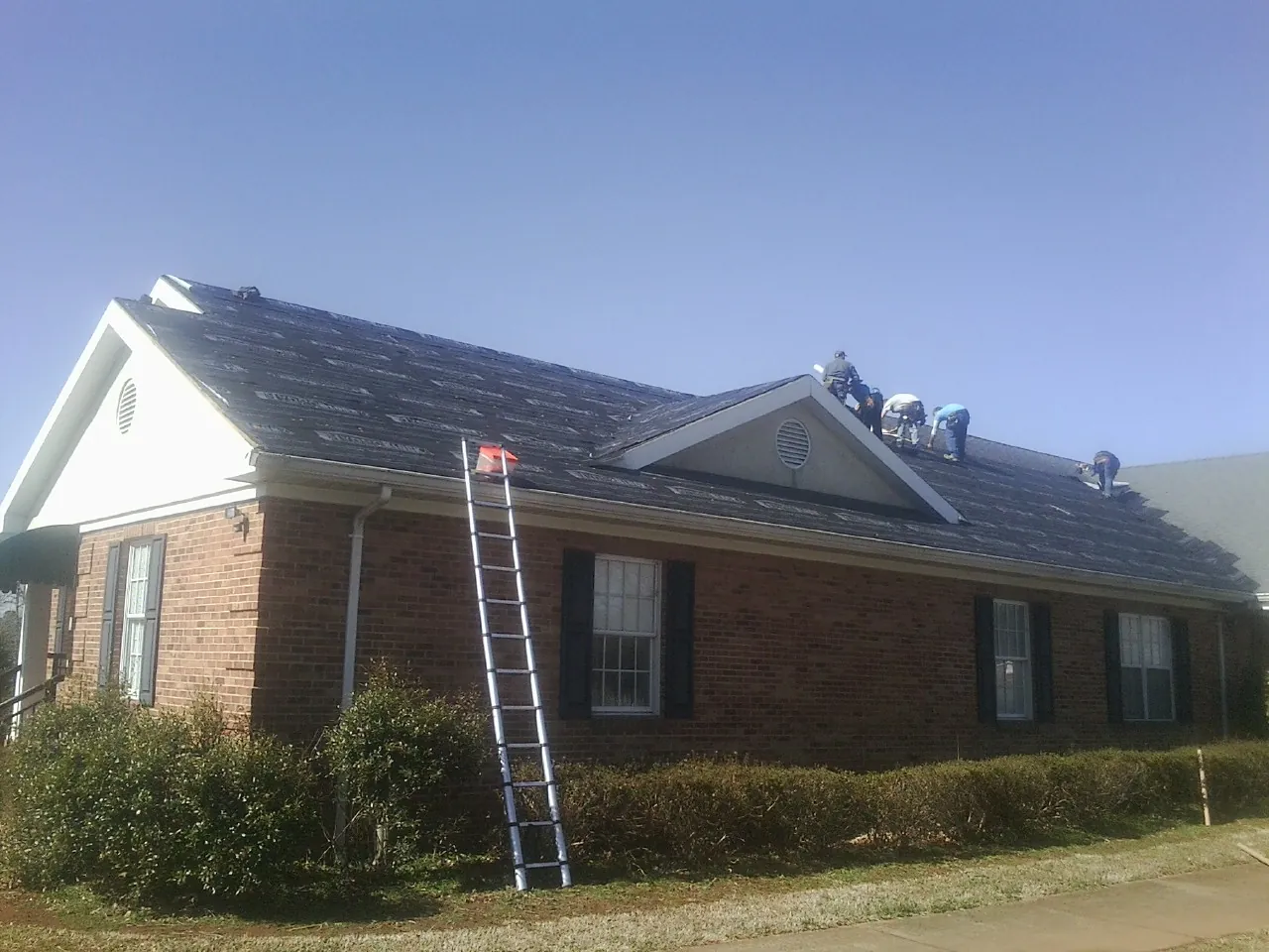 Crew tearing shingles off a brick ranch roof with a ladder set — Metal Roof Repair work by our crew in Gilchrist