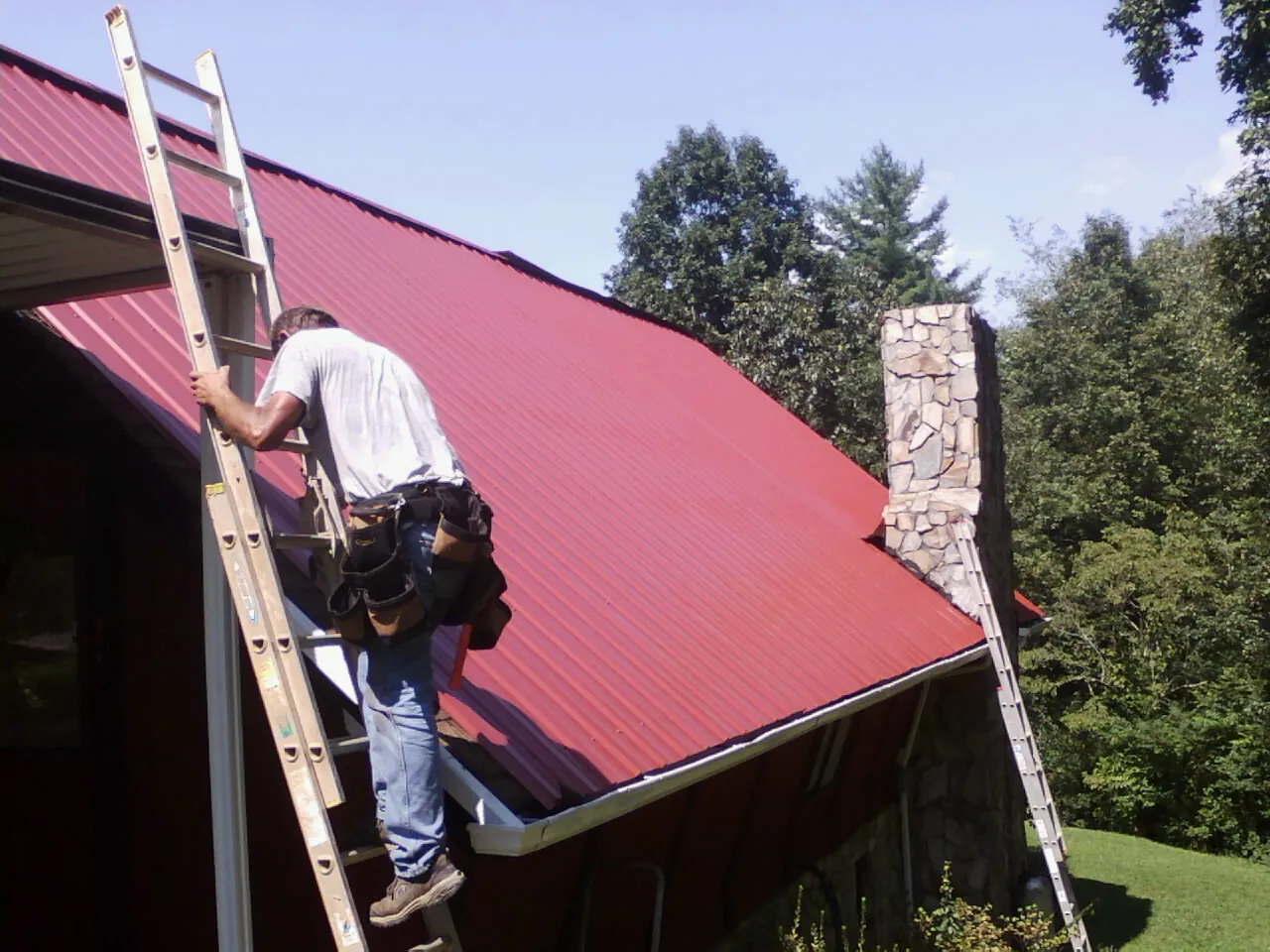Roofer on a ladder beside a red metal roof and stone chimney — Soffit & Fascia Repair by Roofing Brothers in Gilchrist